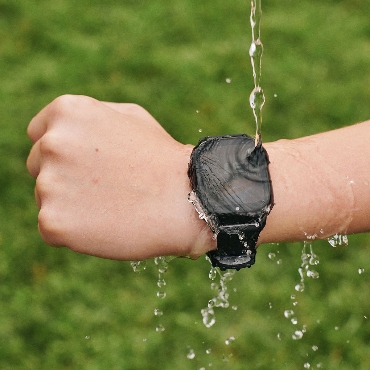 Person's wrist with a black smartwatch under running water against a green grass background