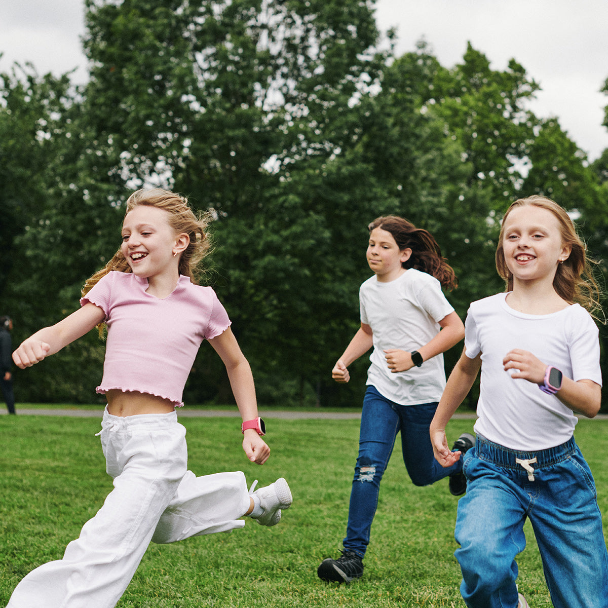 Three children running on a grassy field with trees in the background wearing smartwatches